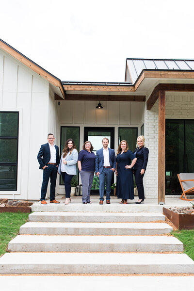 Keeton real estate team are in front of a texan house. there are 6 people in the photo and they wears with elegant clothes and they are looking at the camera.