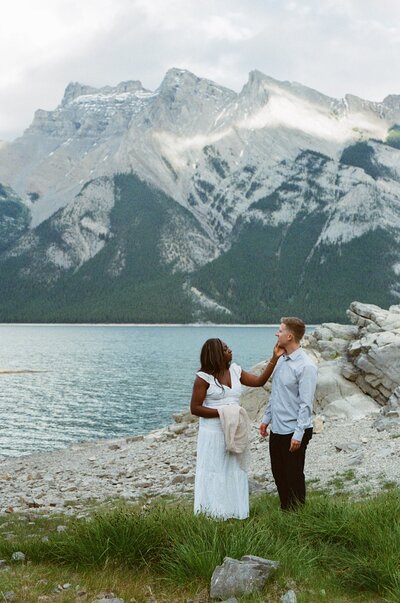 film photo of bride touching groom's face beside Lake Minnewanka in Banff with mountains in the background