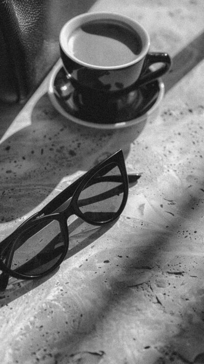 A pair of cat-eye sunglasses and a cup of coffee on a saucer rest on a textured surface, with diagonal shadows across the scene—a touch of Sweet Pea Ink Creative style.