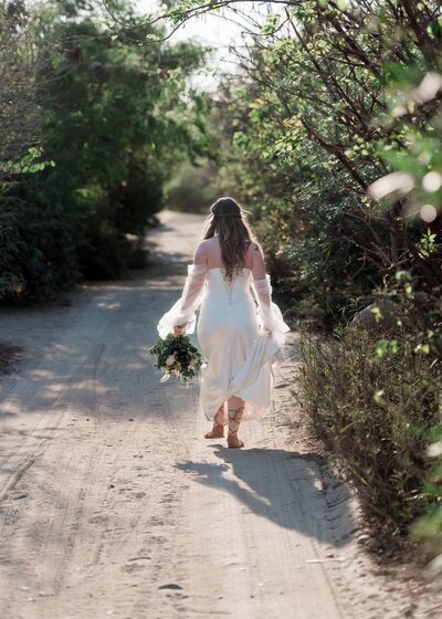 A bride in a beautiful wedding dress walks down a sandy dirt road during her luxury adventure elopement wedding in mexico.  The sun creates a halo around her long hair and glowing lacy sleeves and edges of her dress as she walks away from her elopement photographer. 