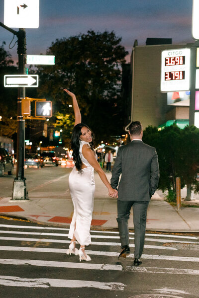 bride and groom crossing a road holding hands while she looks up smiling with her other arm raised up