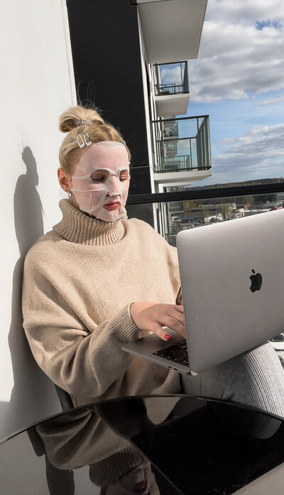 A sunlit home office scene with a coat draped over a chair. A laptop, stacks of books, and a red candle sit on the desk, casting soft shadows.