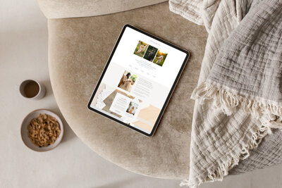 A tablet displaying a website in a day with various images rests on a beige cushion next to a bowl of snacks and a cup of brown liquid, partially covered by a fringed beige blanket.