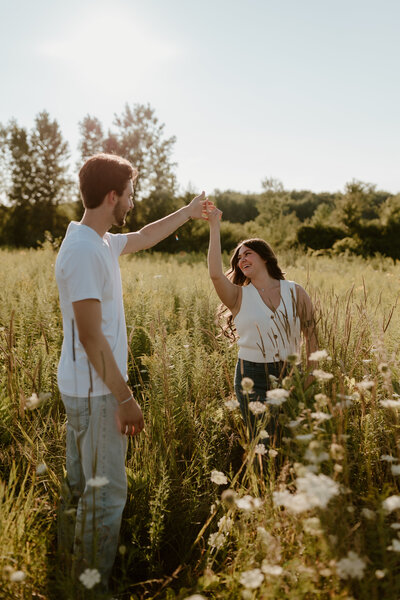 A couple dances in a sunlit field surrounded by tall grass and wildflowers, exuding joy and warmth. The sun casts a soft glow, creating a serene, romantic mood.