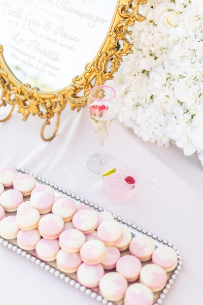Pink and cream macarons on silver tray with champagne rose cocktail and raspberry drink in front of gold mirror menu and white floral wall.