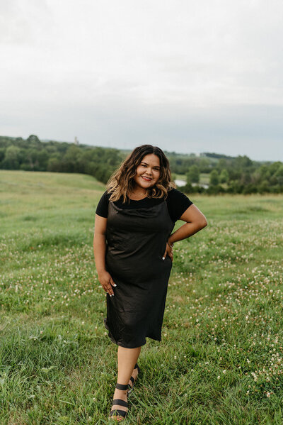 Virginia wedding photographer in a black dress and black shoes posing with a hand on her hip in a spring wild flower field in Shenandoah National Park with rolling hills and mountains in the distance