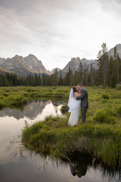 Bride and groom share a kiss in an alpine meadow beside a reflective river, surrounded by towering mountain peaks.