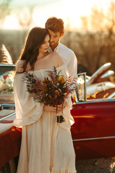 Husband holds his bride from behind while standing in front of a red convertable wearing wedding attire.