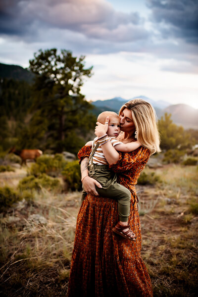 A mother holds her toddler boy and kisses his forehead while the sun peeks through the clouds behind them. 