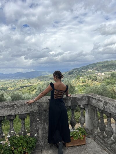 A woman in a black dress stands on a balcony overlooking a sun-drenched Italian winery, embodying timeless elegance amidst the rolling vineyards.