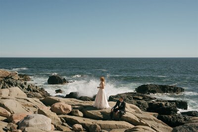 A bride standing looking out at the ocean and a groom sitting looking at the bride overlooking waves at Acadia National Park in Maine. 
