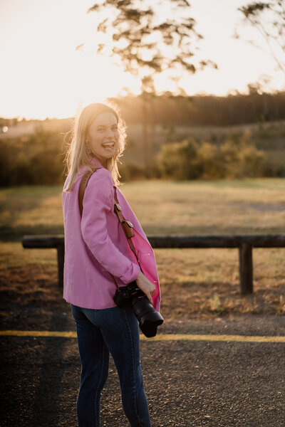 Woman in pink coat with camera over her shoulder looks back at the camera as she walks away - Brisbane wedding photographer
