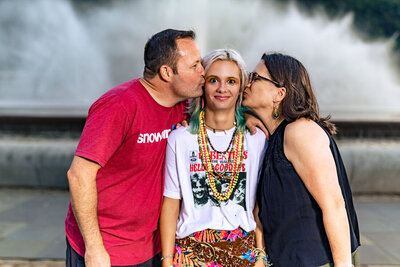 a mother and a father kiss their senior daughter on each cheek and she smiles at the camera with the background of the point state park fountain. 