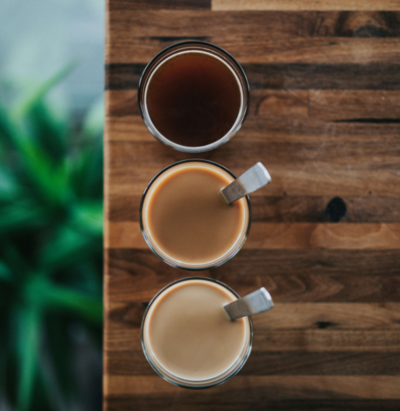 Overhead view of three mugs of coffee or chai in varied shades on a wooden table, representing personalized consulting and inclusive, human-centered experiences.