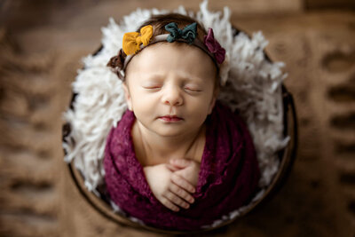 Newborn baby girl wrapped in purple with yellow, green, and purple bows on her headband.