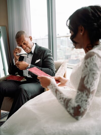 A groom sits in a chair, wiping a tear with a tissue while holding a red card. The bride, in a lace wedding dress, sits in the foreground reading a card.