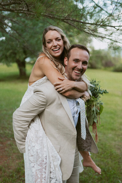 a groom in a tan suit giving a barefoot bride a piggy back ride during their elopement in Vermont