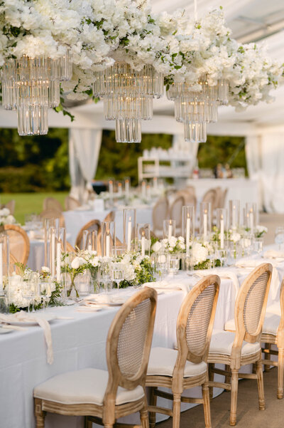 Wedding reception table, white table cloth, wooden cushioned chairs. Above the table are white bouquets and crystal chandeliers