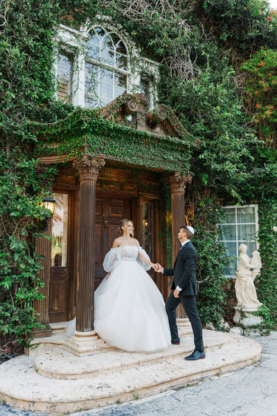 A couple on wedding gowns facing the camera , the groom is sitting down looking to the side while the bride is standing next to him looking at the camera while placing her elbow on his shoulder