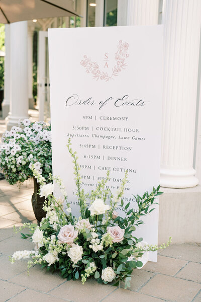 A white wedding event sign displays the order of events, starting at 3 PM with a ceremony and concluding at 7:30 PM with dancing. The sign, courtesy of our full service wedding planner, is adorned with flowers at its base.