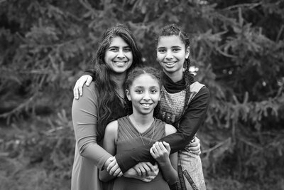 Black-and-white portrait of a smiling mother holding her two daughters, with blurred pine trees in the background at a scenic Kingston location.