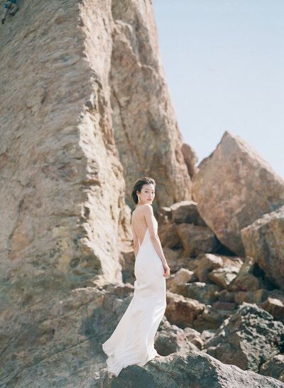 Kohnur modern chic bride with natural subtle makeup and a sleek low bun, wearing an open-back silk slip dress posing on a rocky coastline.