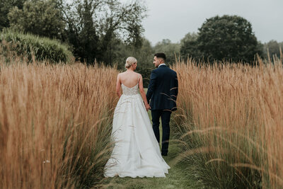 Bride & Groom walking through golden long grass field at Bury Court Barn, Farnham