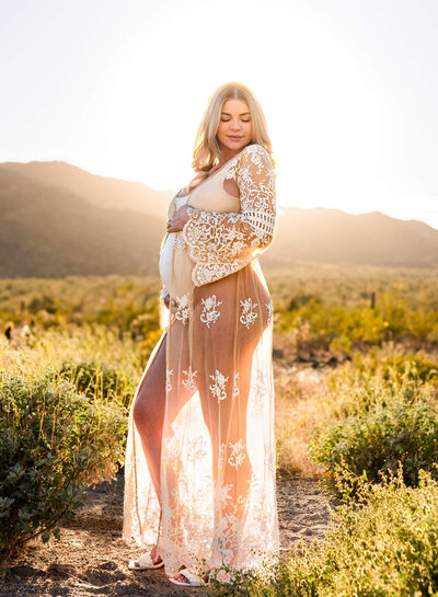 Gorgeous pregnant mom in sheer, white lace dress posing outdoors at sunset
