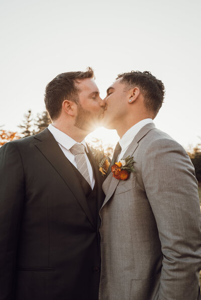 A male couple kisses as the setting sun bursts in between them to create a warm, fall wedding image.