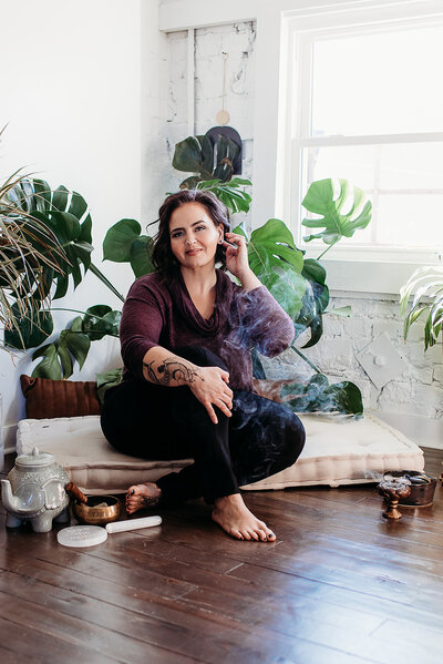 woman is tucking her hair behind her ears while sitting on a large pillow with plants all around her and incense burning beside her