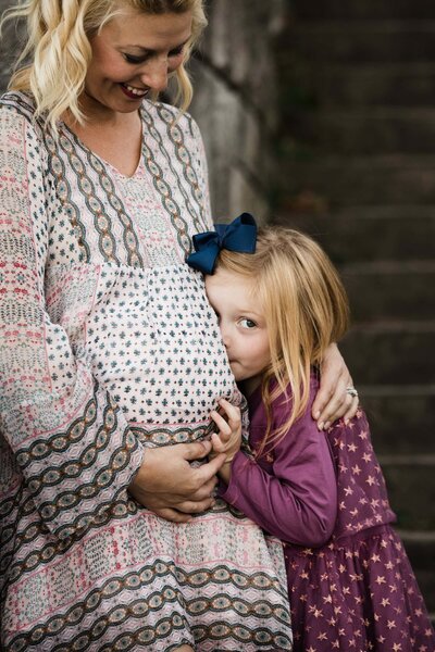 A pregnant mom looks down on her daughter and smiles in a printed dress as her young daughter places her ear to her belly