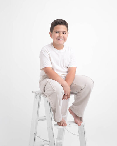 studio portrait of boy on white ladder with white backdrop wearing white shirt and light beige pants