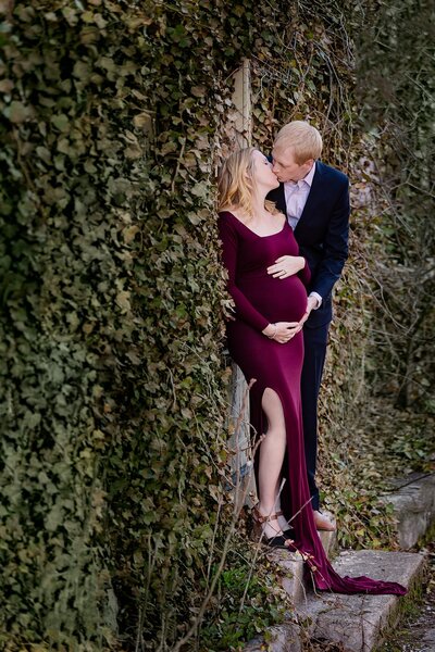 expactant parents kiss standing near wall of ivy