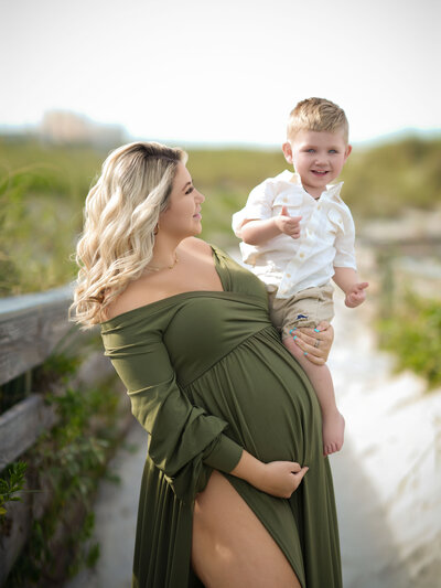 A pregnant woman in a flowing green dress holds a smiling child on a sandy path flanked by greenery, conveying warmth and joy on a sunny day.