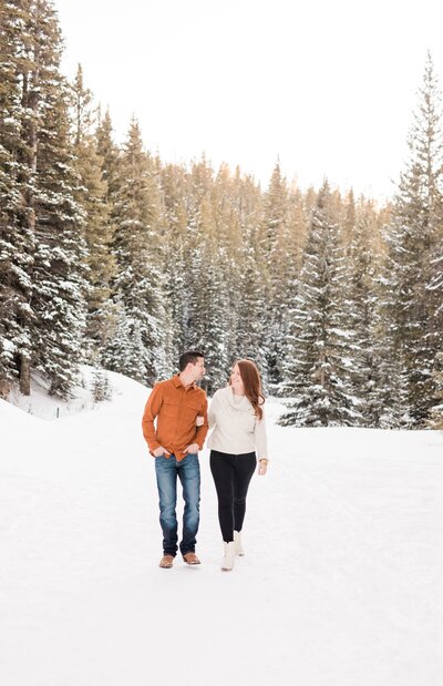 A happy couple walks arm in arm in a snowy Boreas Pass trail at sunset smiling at each other
