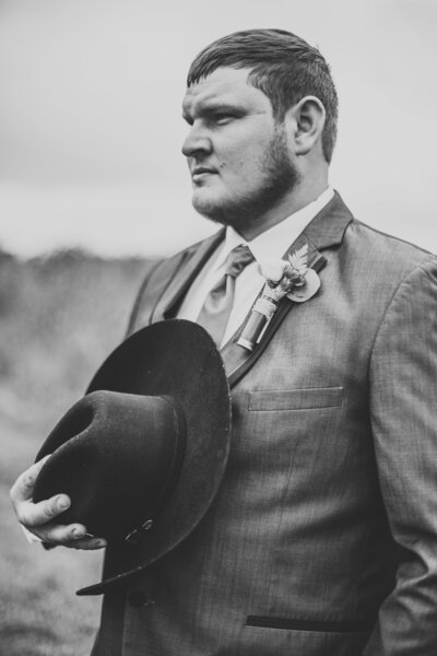 Black and white photo of cowboy groom holding cowboy hat on chest gazing into the distance by Iowa City wedding photographer Sabrina Wilham