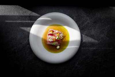 Zwarte ondergrond met bord waarop een heerlijk lente gerecht van hamachi, biet, bloemen, dashi. Kleurrijk gerecht door inval zonlicht. © Studio Ensō