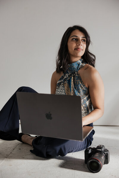A person with long dark hair sits on the floor with a laptop on their lap, looking to the side. They wear a patterned sleeveless top and dark pants. A camera rests nearby, hinting at their passion as a New England Lifestyle Photographer. The background is plain white.
