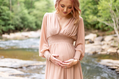 Maternity portrait outdoors by a creek with mom to be wearing light pink dress holding both hands on bottom of baby bump and looking down at tummy