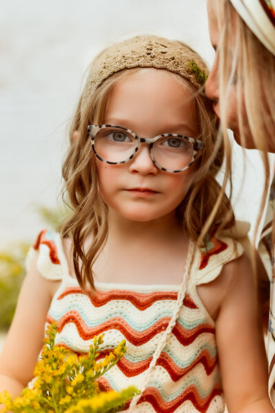 Sweet girl in glasses with curly blonde hair and a purse full of wildflowers stares at the camera in her Kansas City Family Photoshoot.