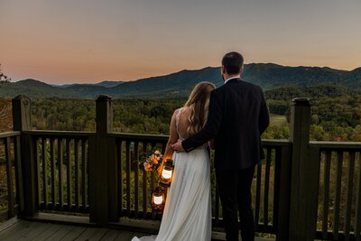 A bride and groom stand looking out at the sun set behind a vast mountain range after eloping. The groom has his arm around the bride while she leans her head on him. The sky is pink, blue and orange from the sunset while the vast mountain range is a mix of blue and green. They are both in wedding gear holding lanterns.