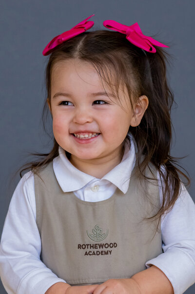 Girl in school unirform and pink hair bows looing off to the side but still smiling for her school photos