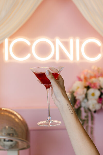 Hand holding a cocktail glass in front of a neon “ICONIC” sign and pink floral salon scene — detail image from a vibrant portrait shoot by Greenville South Carolina portrait photographer Dani Lacey Photographs.