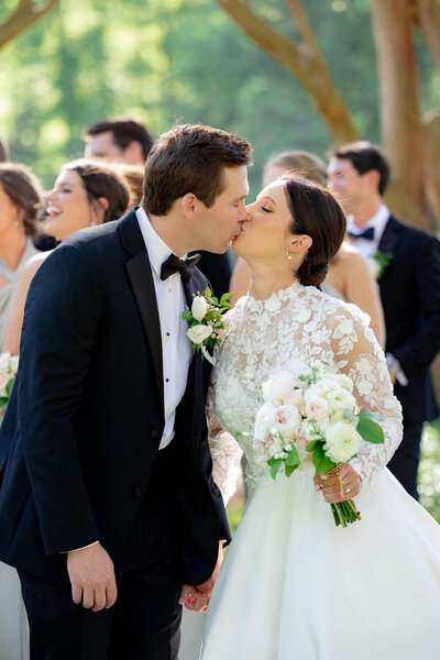 man in black tux kissing woman in white dress holding a bouquet of flowers