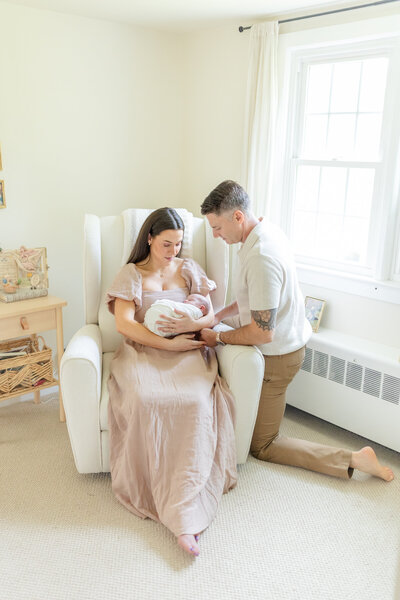 Newborn session photo in the client's home nursery, where mom sits in a rocker holding the baby, while dad kneels beside them, lovingly looking at their newborn. The warm, cozy nursery setting adds to the intimate family moment
