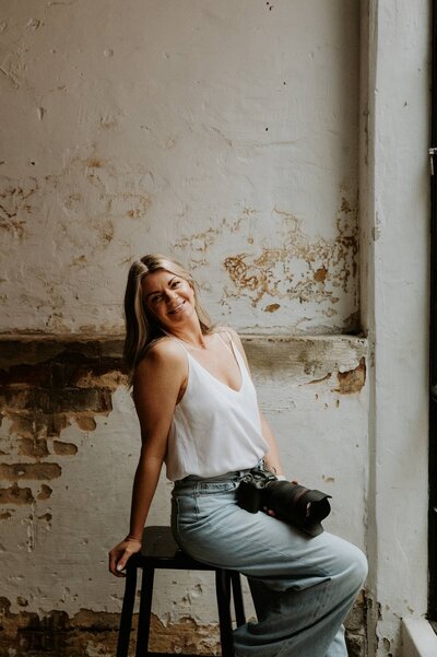 Kaz Soom, Perth Maternity, Newborn and Family Photographer.  She is sitting on a white folding chair, holding her camera and smiling at the camera. 