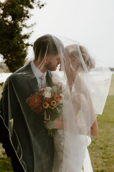 A bride and groom under a delicate veil share an intimate moment outdoors. The bride holds a bouquet with vibrant flowers, evoking a sense of romance and joy.