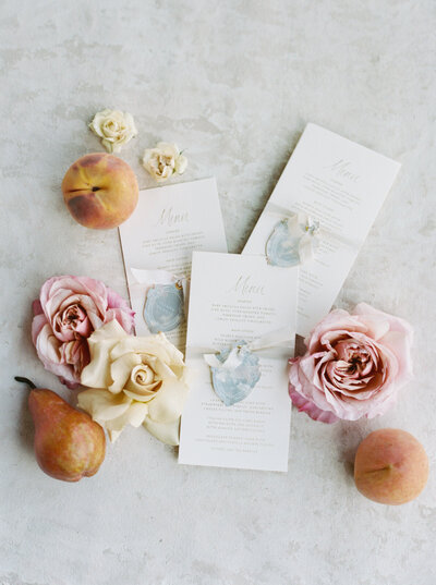 High-end wedding stationery items styled on a flat art surface, surrounded by roses and fresh fruit