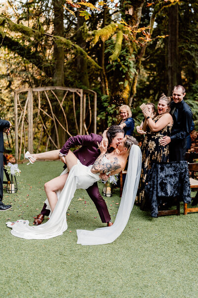LGBTQ+ seattle wedding couple dip kiss at forest wedding with guests cheering