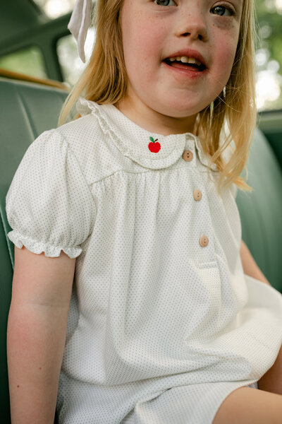 A little girl wearing and white and green sage dot knit dress with ruffle sleeves and buttons and red embroidered apples back to school